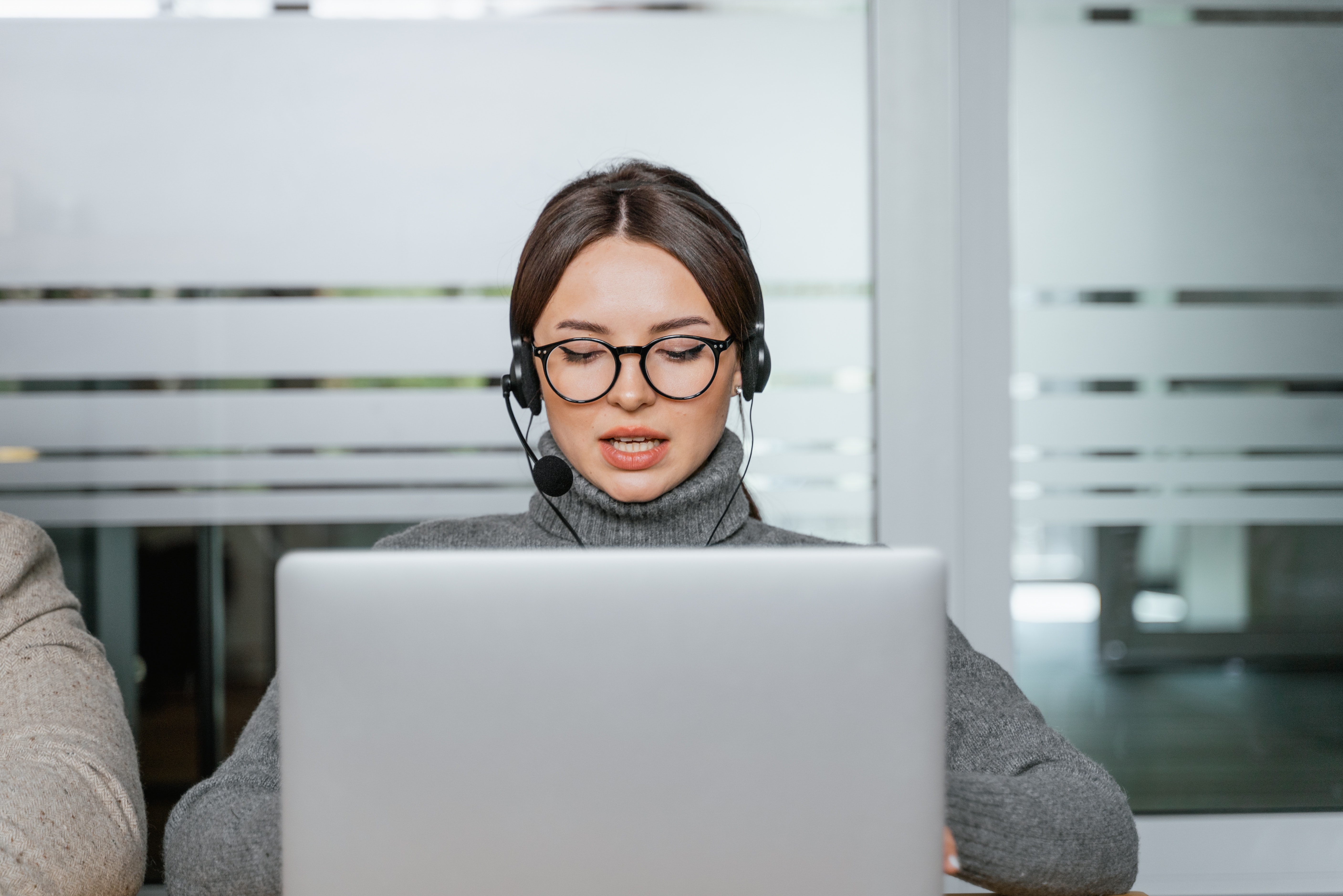 mlada žena smeđe kose u naočalama sjedi za laptop sa slušalicama u ušima i mikrofonom