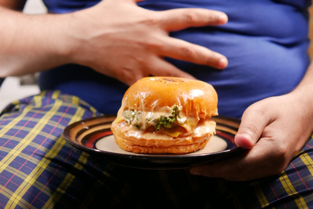 čovjek s velćim trbuhom i hamburgerom na tanjuru ispred sebe