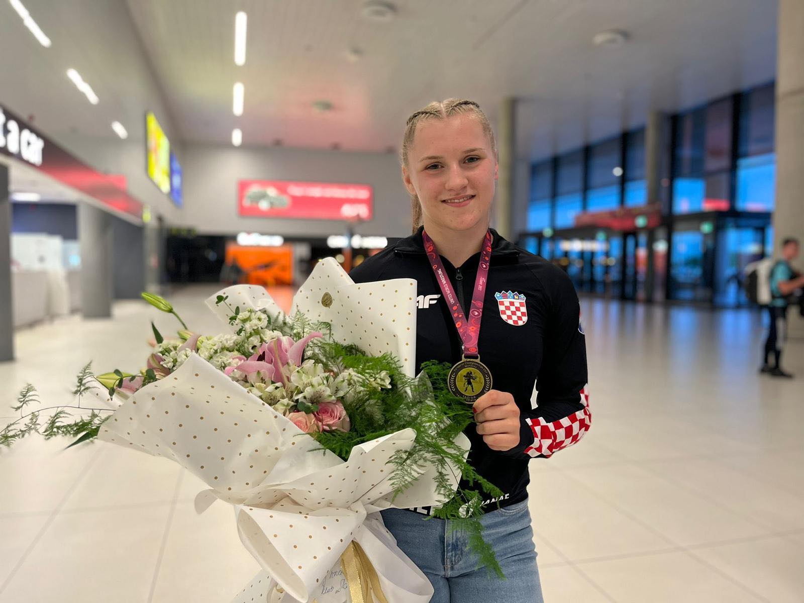 veronika vilk, europska prvakinja u hrvanju, prikazana je na slici s buketom cvijeća, zlatnom medaljom u rukama te jaknom s hrvatskim 'kockicama'