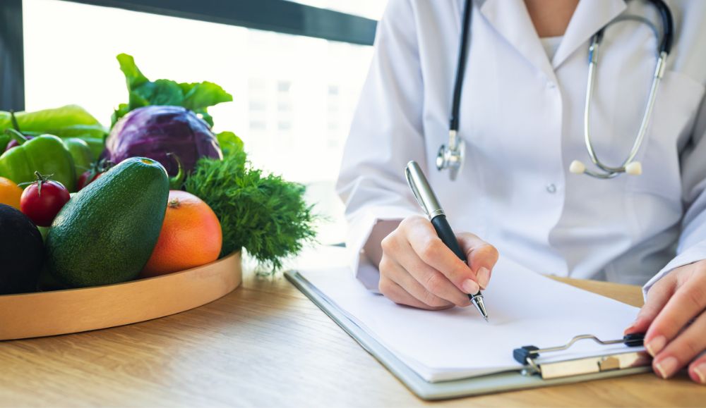 nutricionist u kuti sa stetoskopom za stolom piše nešto po papiru, na stolu je zdjela s povrćem i voćem